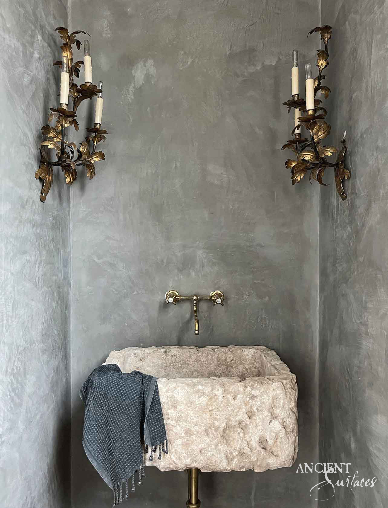 Reclaimed antique limestone trough sink from Ancient Surfaces with a rough hewn quarry face, set in a hand troweled plaster alcove between gilt candle sconces