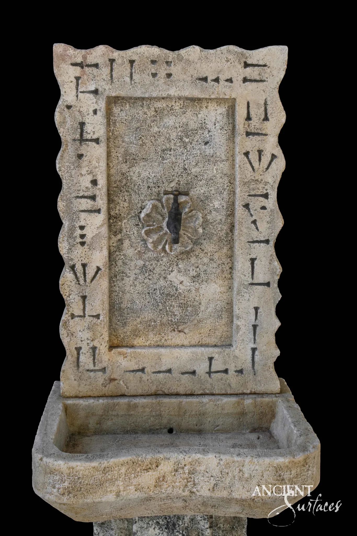 Reclaimed antique limestone wall panel sink from Ancient Surfaces with an incised geometric border and a carved rosette spout fitting, photographed against black