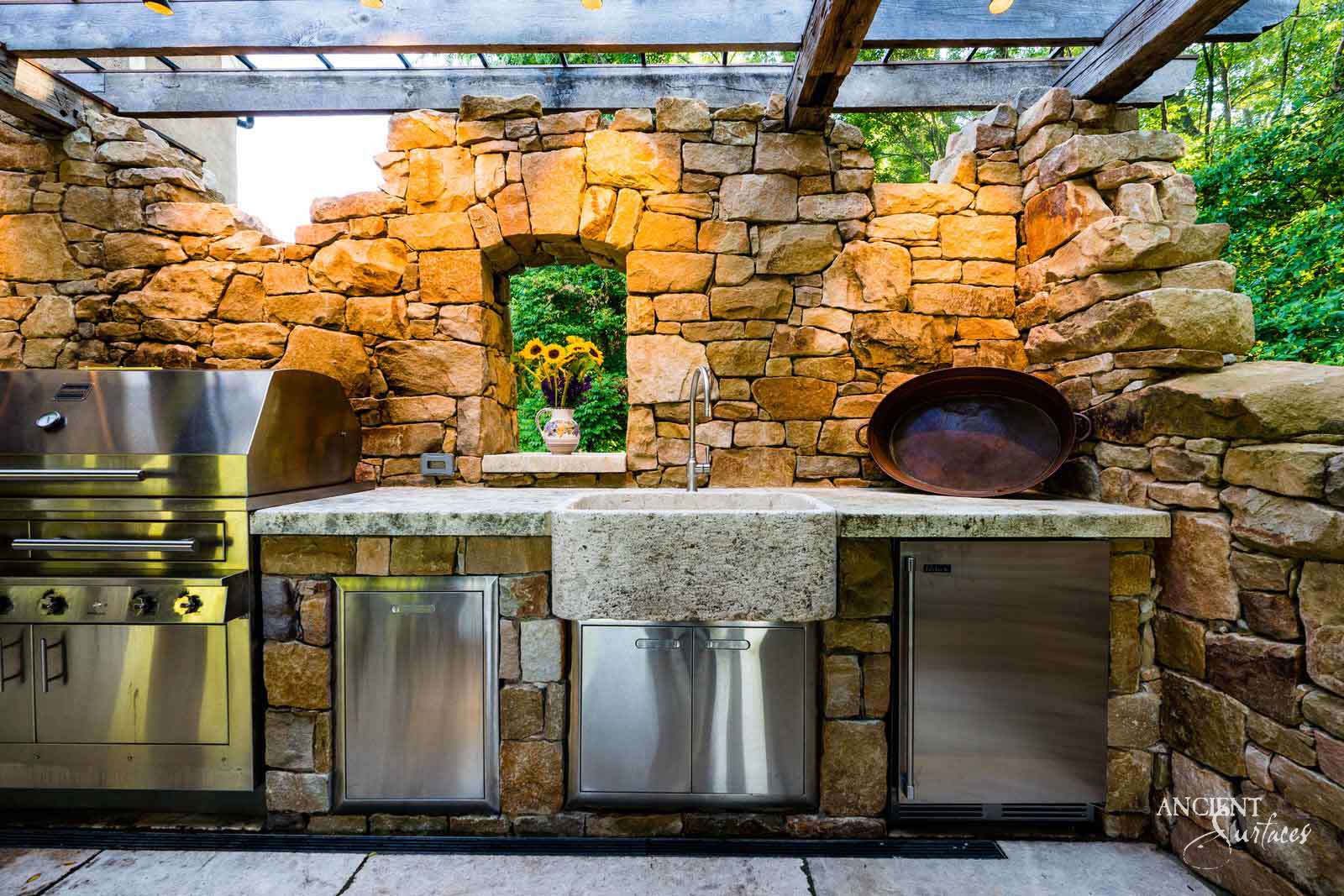 Reclaimed antique stone sink from Ancient Surfaces set into a granite counter within an outdoor fieldstone kitchen, between a grill and refrigeration drawers