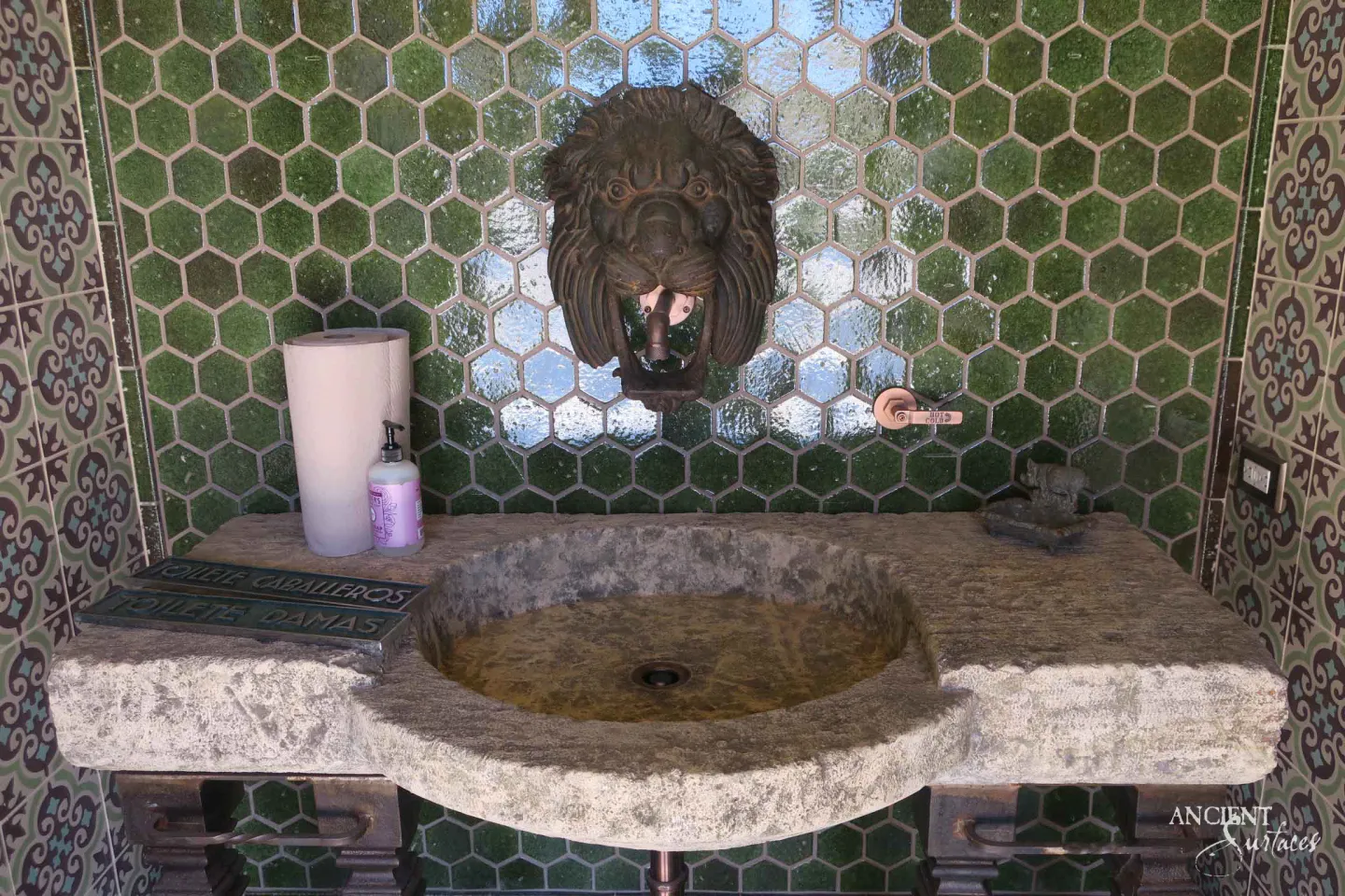 Reclaimed antique limestone sink from Ancient Surfaces with a carved oval basin beneath a cast bronze lion mask spout, mounted on a green hexagonal tile wall