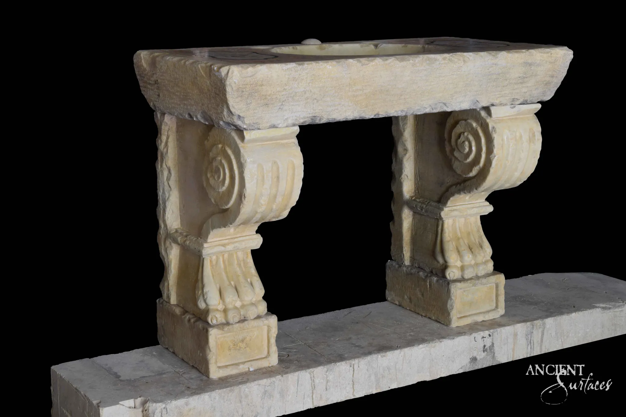 Reclaimed antique limestone bench style sink from Ancient Surfaces with a shallow carved basin on two scrolled volute corbel legs, photographed against black