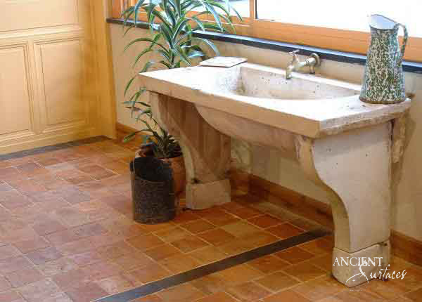Reclaimed antique limestone trough sink from Ancient Surfaces on shaped corbel legs, installed in a terracotta floored utility room beside a potted palm