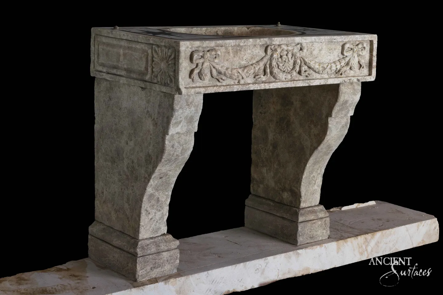 Reclaimed antique limestone console sink from Ancient Surfaces carved with a rosette and ribbon festoon swag, with a dark inset stone top, photographed against black