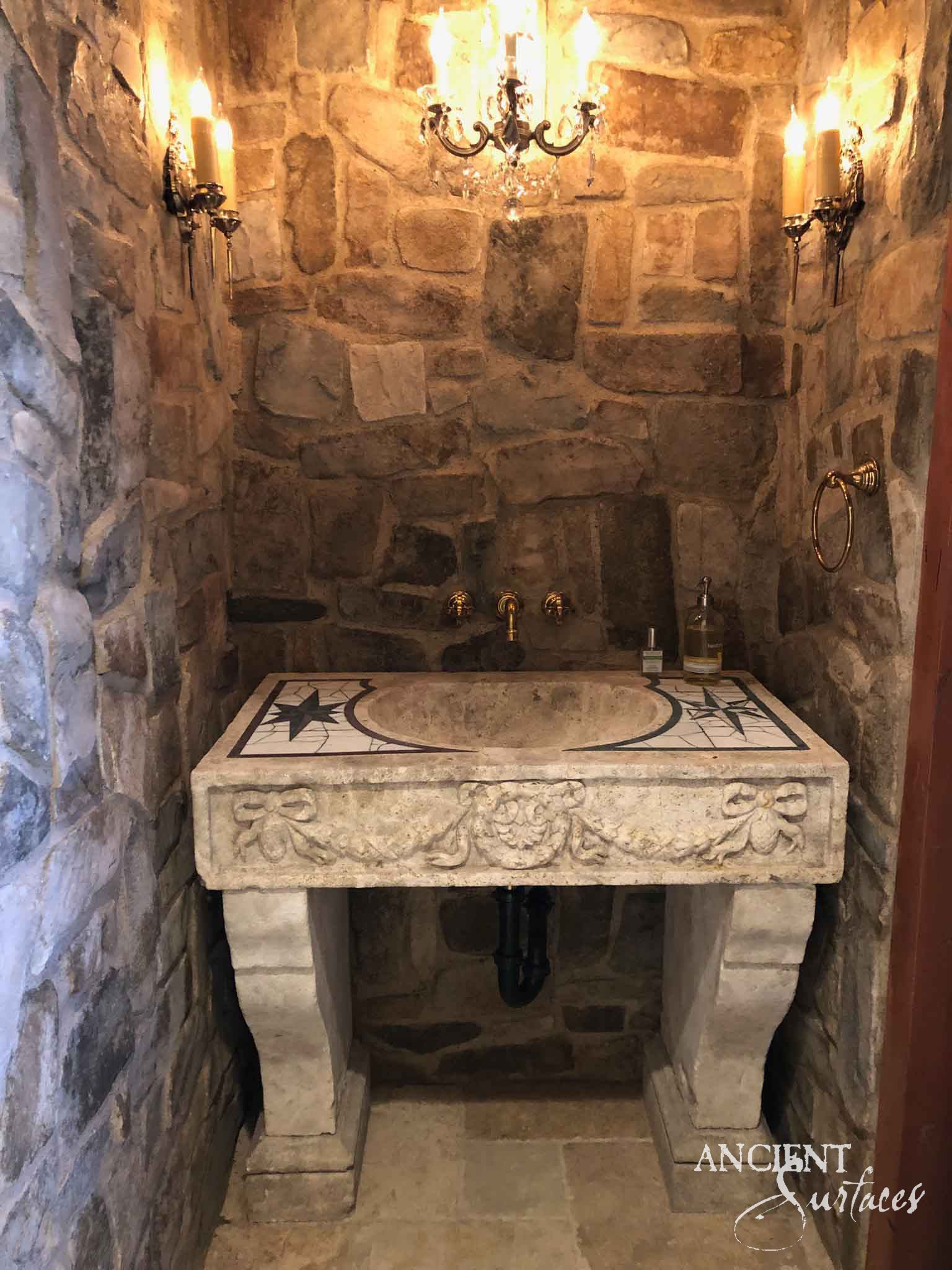 Reclaimed antique limestone vessel sink from Ancient Surfaces on a stone plinth inside a rough stone grotto bathroom lit by a small chandelier and wall sconces